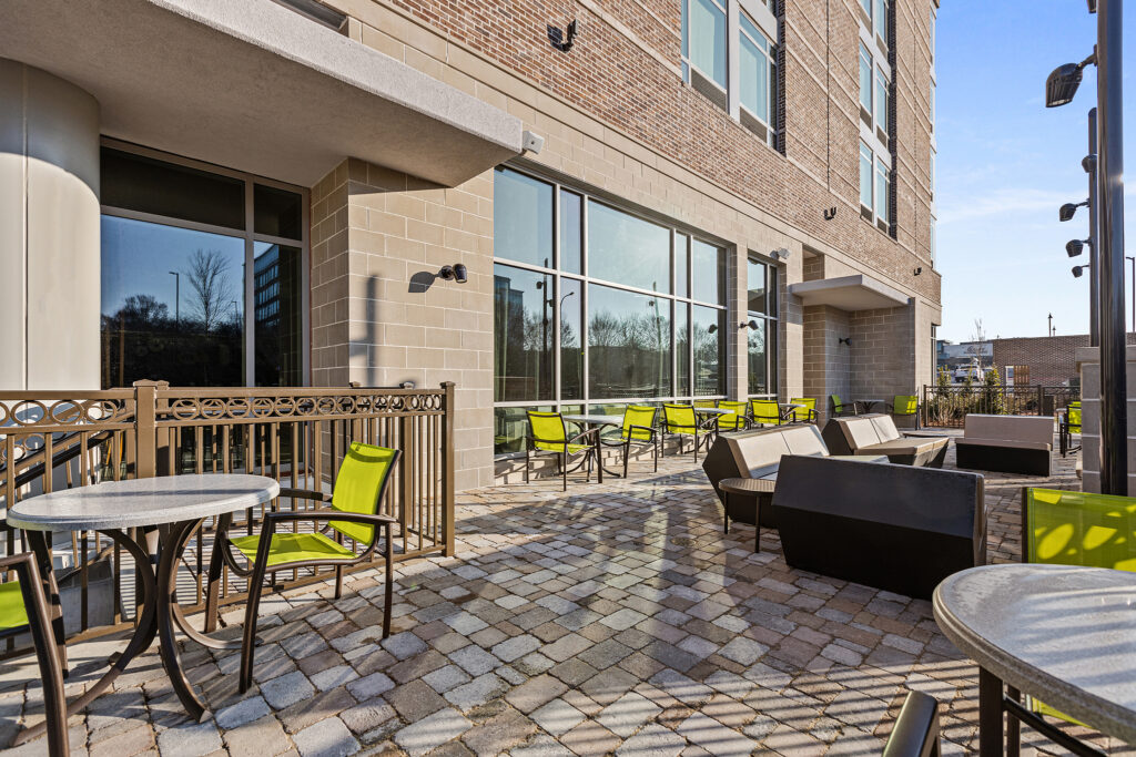 The outdoor patio area features modern stone flooring with metal tables and green chairs, perfect for guests at the SpringHill Suites in Coolsprings, TN. Adjacent to a brick building with large glass windows, it's a serene spot beneath the clear blue sky.