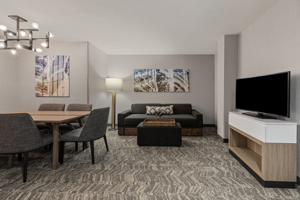 A modern living room at SpringHill Suites in Coolsprings, TN, features a gray sofa, patterned rug, and two abstract paintings. A wooden coffee table and matching dining table with chairs sit under a unique chandelier. A TV is on a white stand, and a floor lamp stands in the corner.