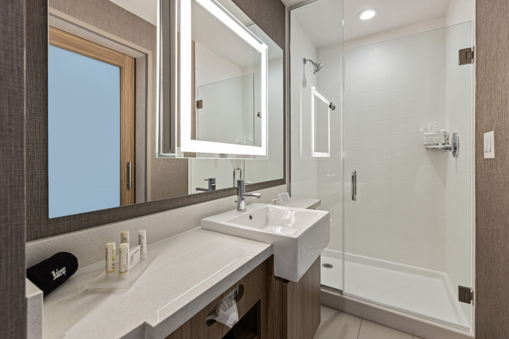 A modern bathroom at SpringHill Suites Coolsprings TN features a large illuminated mirror above a rectangular sink. The glass-enclosed shower and neatly arranged toiletries embellish the light-tiled walls, creating a tranquil retreat within this Marriott property.