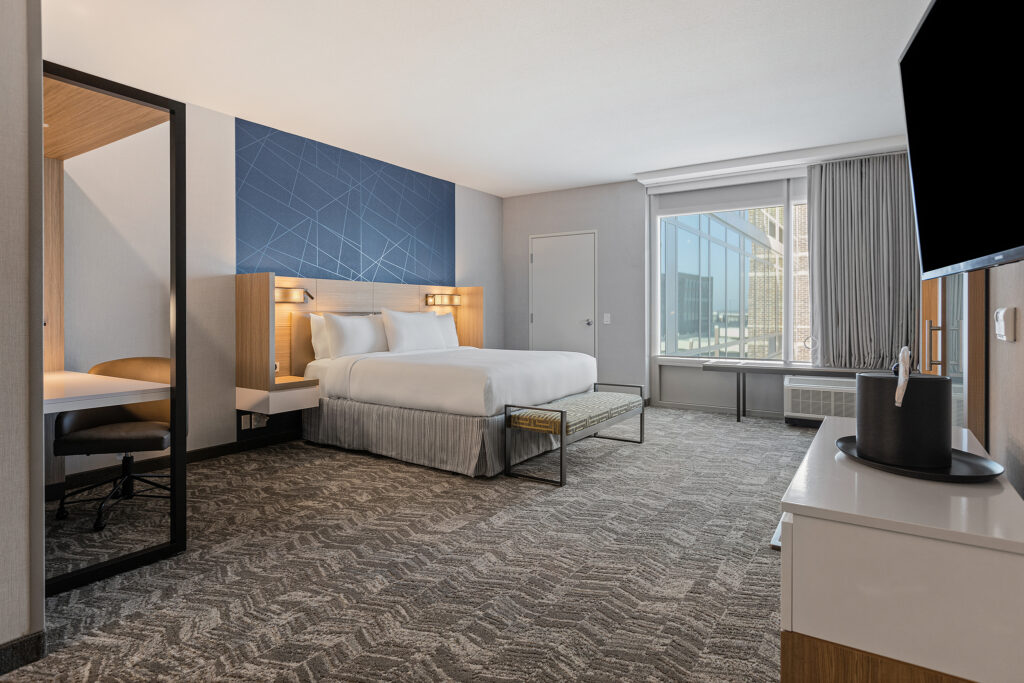 A modern hotel room at SpringHill Suites Marriott in Cool Springs, TN, featuring a large bed with white linens, a blue accent wall, a desk with a chair, and a flat-screen TV. The window offers captivating city views. The decor is minimalistic with neutral tones and clean lines.