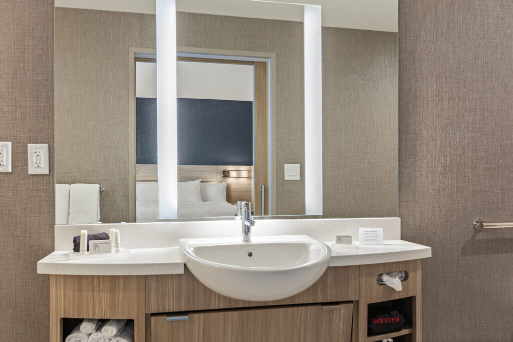 The modern bathroom at SpringHill Suites in Coolsprings, TN boasts a sleek vanity with a round white sink and a lighted mirror. Towels, toiletries, and a tissue box are neatly arranged on the counter. The textured beige wall adds warmth, with a glimpse of the bed through the doorway.