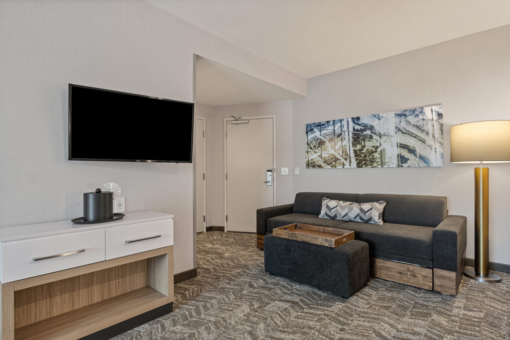 This modern Marriott hotel room in Coolsprings, TN, features a wall-mounted TV, a gray sofa with patterned cushions, a wooden coffee table, and a standing lamp. Abstract art adorns the walls of this SpringHill Suites gem, with its soothing neutral palette. There's also a door in the background.