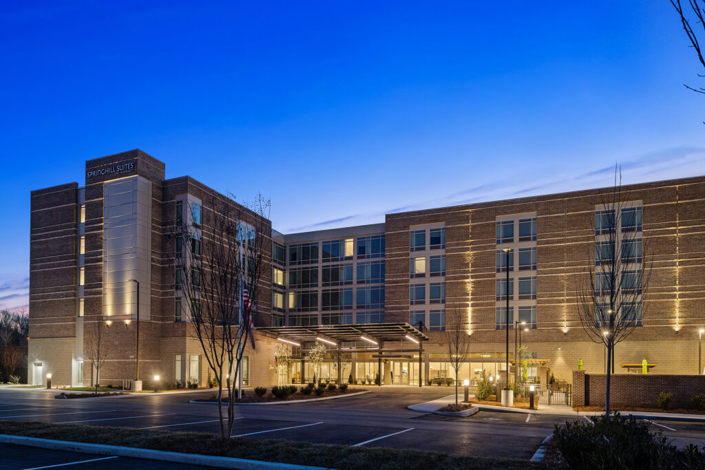 A modern hotel building at dusk with illuminated windows and a well-lit entrance, this SpringHill Suites by Marriott captures the charm of Coolsprings TN. The facade features brick and glass elements against a gradient blue sky as evening sets in, with a few bare trees in the foreground.