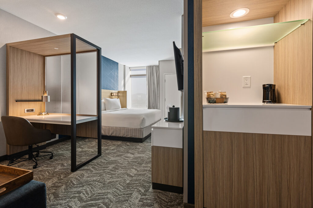 Modern hotel room at SpringHill Suites in CoolSprings, TN, featuring a queen-sized bed, a work desk with chair, and a wall-mounted TV. To the right, a kitchenette area includes a coffee maker. The room is decorated in neutral tones with contemporary design synonymous with Marriott's standards.