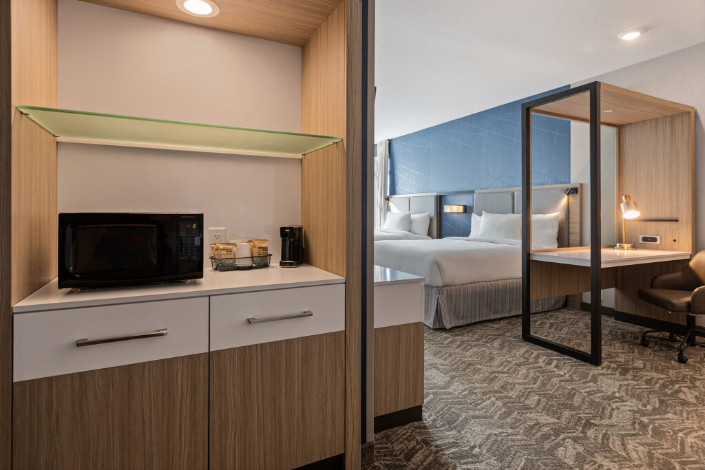 This SpringHill Suites hotel room in CoolSprings, TN, boasts a modern design. The foreground features a kitchenette with a microwave, coffee maker, and drawers. In the background, there's a bed with white linens, alongside a desk with a lamp and chair on patterned carpet.