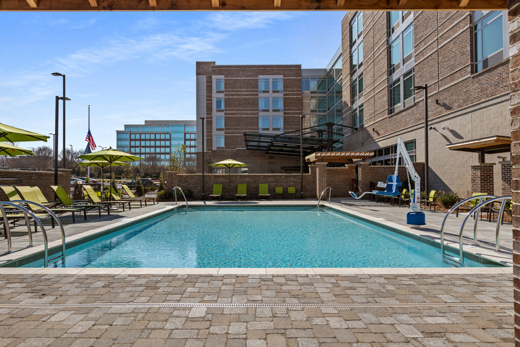 The outdoor swimming pool at SpringHill Suites in Coolsprings, TN, boasts clear blue water surrounded by lounge chairs and green umbrellas, next to a modern brick building. A lift for accessibility is visible poolside, with an American flag waving under the sunny sky.