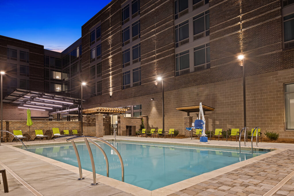A well-lit outdoor pool area at night at SpringHill Suites Coolsprings TN, surrounded by a brick building. Green deck chairs are placed around the pool, and lights illuminate the area. A small pergola and pool lift enhance this Marriott property's inviting ambiance.