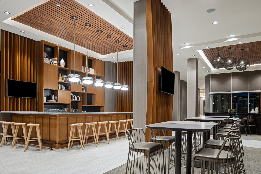 A modern interior at SpringHill Suites Coolsprings TN features a long wooden bar with stools, pendant lights, and shelves. Adjacent are tables and chairs. The space boasts a minimalist design with wooden elements, neutral tones, and a contemporary ceiling detail that embodies Marriott elegance.