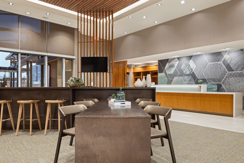 Modern office space at SpringHill Suites in Coolsprings, TN, featuring a long wooden table and chairs, a high counter with stools, geometric wall art, and a decorative wooden partition. Large windows provide natural light to this sleek and professional Marriott environment.