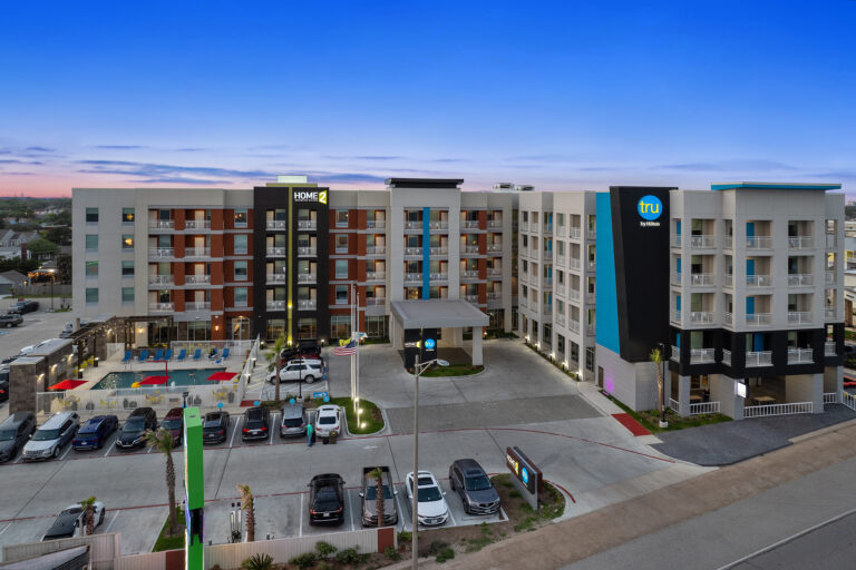 A modern hotel complex at dusk showcases two interconnected buildings. The left proudly displays the Home2 by Hilton logo, while the right features Tru. Captured by a drone, colorful lights illuminate the facilities, with a parking lot visible in the foreground.