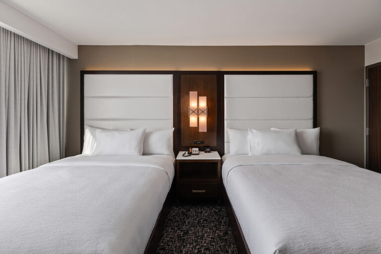 A room at Embassy Suites in Syracuse offers two neatly made double beds with crisp white linens. Between the beds, a wooden nightstand with a modern lamp stands, while floor-to-ceiling curtains grace the window on the left. The cozy ambiance is completed by carpeted flooring.