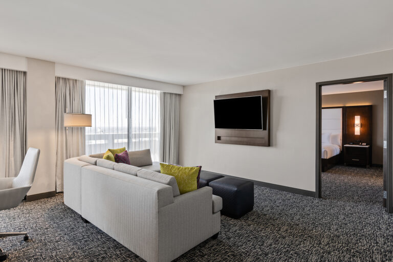 This modern Hilton hotel suite in New York features a neutral color palette. The living area has a light gray sofa with colorful cushions, a wall-mounted TV, and a window with sheer curtains. An adjacent bedroom is partially visible through an open doorway.