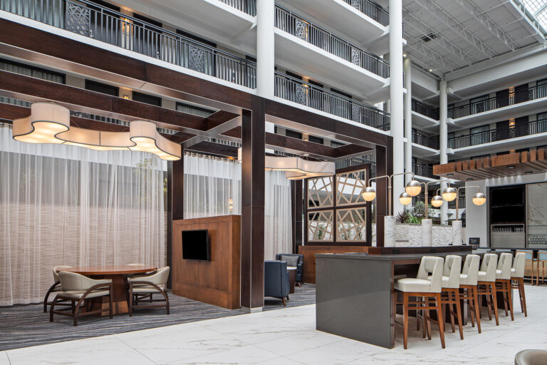 The spacious modern lobby of this Syracuse hotel boasts high ceilings and abundant natural light. It features a seating area with a round table, cushioned chairs, and a high-top bar with barstools. Contemporary light fixtures and wood accents add warmth to the inviting New York space.