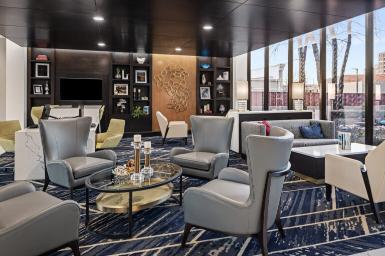 The modern and stylish lounge area at the Embassy Suites in Oklahoma City features gray armchairs, a glass coffee table, and abstract wall art. Large windows let in natural light, while shelves hold decorative items. The space boasts a dark wood ceiling and a blue patterned carpet.