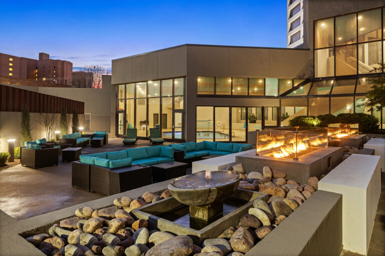 The outdoor lounge area at dusk features modern wicker sofas with teal cushions, creating a perfect retreat at the Hilton. A stone water fountain and glass fireplace enhance the ambiance, while large windows offer glimpses indoors and urban buildings silhouette against the Oklahoma skyline.