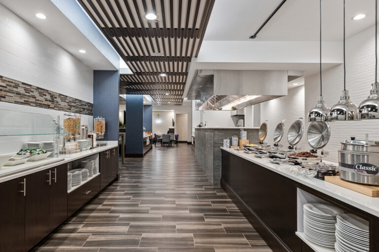 A modern buffet restaurant in the heart of Syracuse, featuring sleek, dark wood flooring and white walls. The buffet line at this Hilton venue offers plates, bowls, cereals, and a variety of dishes under silver heat lamps. Wooden slats grace the ceiling with tables visible in the backdrop.