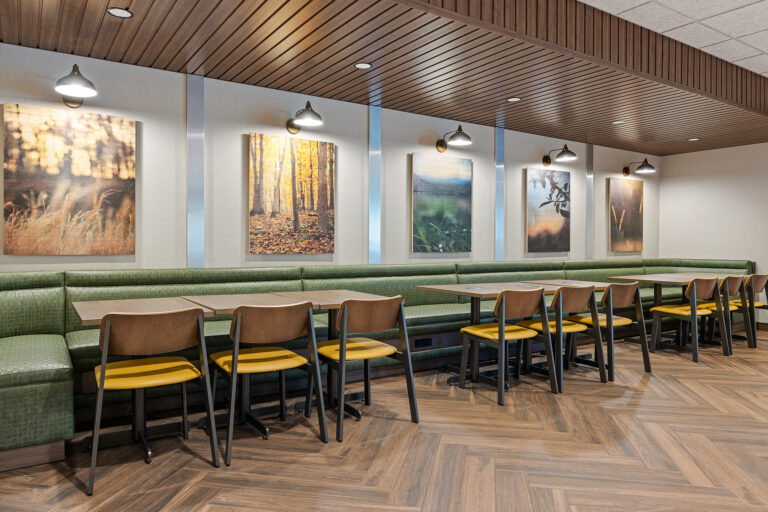 A modern café interior at the Fairfield Inn, featuring green cushioned benches, wooden tables, and yellow-accented chairs. Nature-themed artwork adorns the walls under soft lighting. The herringbone wood pattern floor and ceiling with wood panels create a cozy atmosphere perfect for hotel photography.
