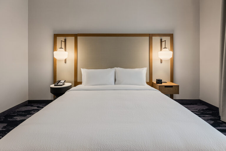 A modern Fairfield Inn hotel room boasts a neatly made double bed with a sleek headboard featuring mounted lamps on either side. Small nightstands hold a telephone and alarm clock, enhancing the minimalist design captured beautifully in hotel photography, all set in a neutral color palette.