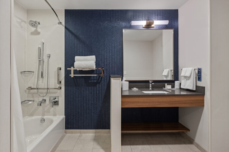 Experience the elegance of a Marriott hotel bathroom with its modern design. Featuring a tub, handheld shower, and secure grab bars, it boasts wall-mounted shelves and a spacious vanity. The dark blue accent wall adds to the minimalist aesthetic, perfect for stunning hotel photography.