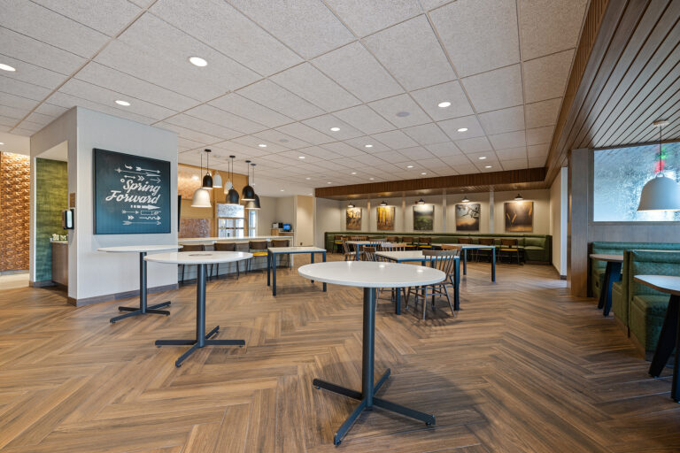 A spacious modern café within the Fairfield Inn features wooden herringbone flooring, round tables, and chairs. Pendant lights hang from the ceiling above. A chalkboard sign reads Spring Forward, and a row of booths lines one wall, creating a Marriott ambiance that is warm and inviting.