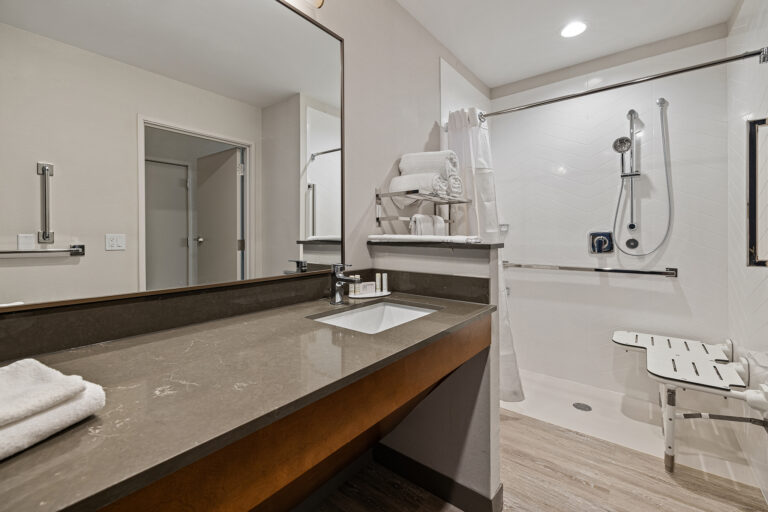 Modern bathroom at the Fairfield Inn features a large mirror, sink on a polished countertop, and neatly stacked towels. The shower area boasts a sliding door, white tiles, and a foldable seat. Light-colored walls enhance the brightness, reflecting the hotel's commitment to comfort and style.