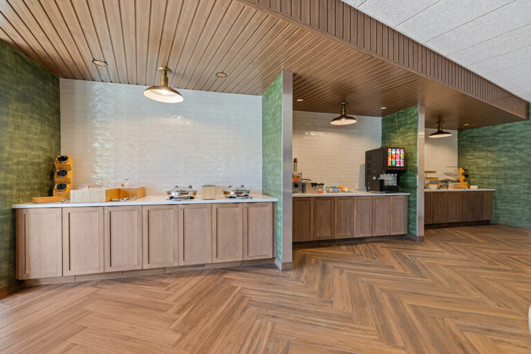 A modern dining area at the Marriott with wooden cabinets and green walls features a self-serve breakfast station. It includes cereal dispensers, a waffle maker, a beverage machine, and various condiments on white countertops. The floor displays a charming herringbone pattern.
