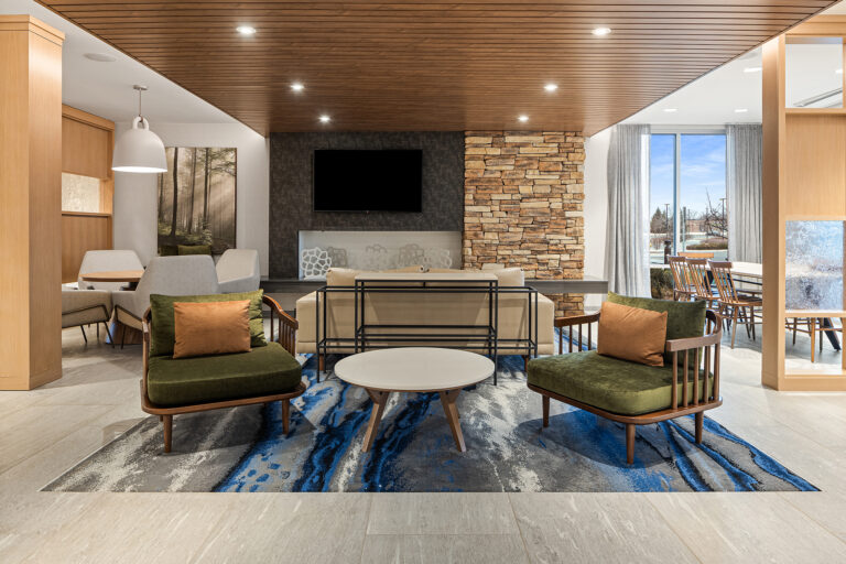 A modern living room, reminiscent of hotel photography, features green armchairs and a beige sofa around a round coffee table on a blue and gray rug. The stone wall with fireplace, wooden ceiling, and large windows offering outdoor views evoke the comfort of a Marriott or Fairfield Inn setting.