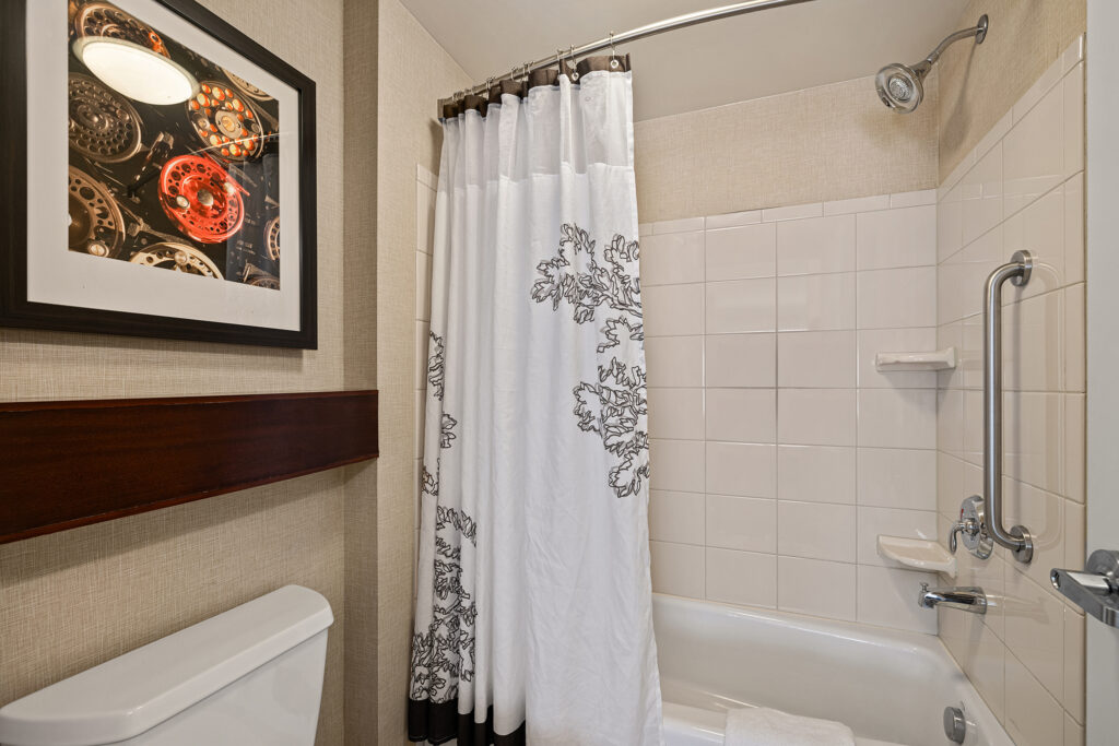 The bathroom at Marriott Glenwood Springs, Colorado, features a white bathtub with a floral-patterned shower curtain, tile walls, and a chrome showerhead. A toilet is beside the tub, complemented by a framed picture of vintage clock gears on the wall.