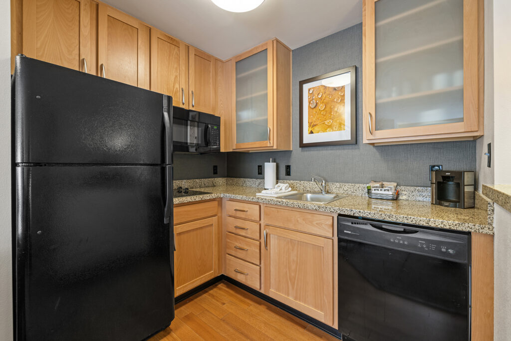 Situated in a cozy Residence Inn, this compact kitchen in Glenwood Springs, Colorado, boasts wooden cabinets, black appliances, and a granite countertop. It features essential amenities like a refrigerator, dishwasher, microwave, and coffee maker. A framed artwork graces the wall above the sink on the hardwood floor.