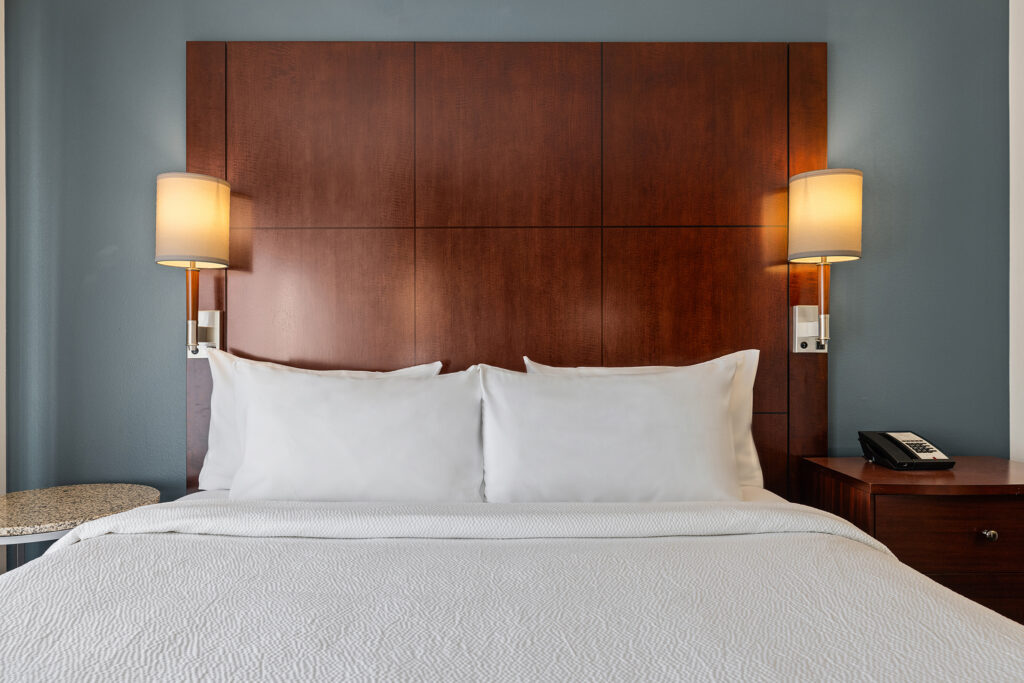 A neatly made hotel bed with white linens and two pillows against a dark wooden headboard greets you at the Residence Inn by Marriott. The blue-painted wall features dual wall-mounted lamps, and a bedside table with a phone sits to the right, capturing the serene essence of Colorado.