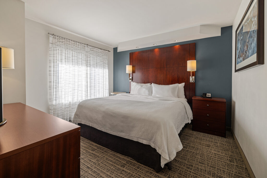 A modern hotel room at the Residence Inn in Glenwood Springs, Colorado, boasts a large bed with white bedding and a wooden headboard. Two wall-mounted lamps offer light. A window with sheer curtains is on the left, while a dresser and artwork on the wall complete this inviting space.