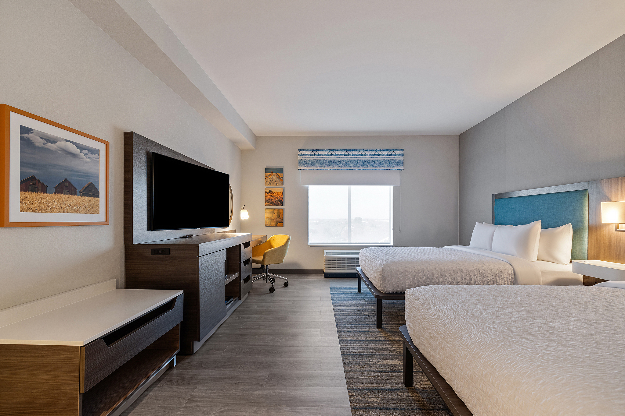 Hampton Inn (4) A modern Hilton hotel room with two double beds, a large flat-screen TV, a desk with a chair, and a window. Neutral tones and contemporary decor are featured, with artwork on the walls and a striped rug on the floor, perfect for stunning hotel photography.
