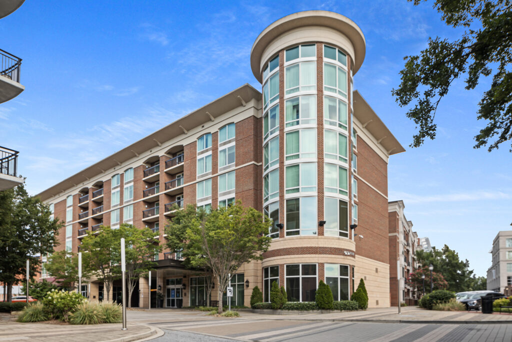 A modern, multi-story brick and glass apartment building with a cylindrical corner resembles the sleek design often associated with Hilton hotels. It boasts a well-maintained entrance and surrounding trees under a clear blue Greenville sky.