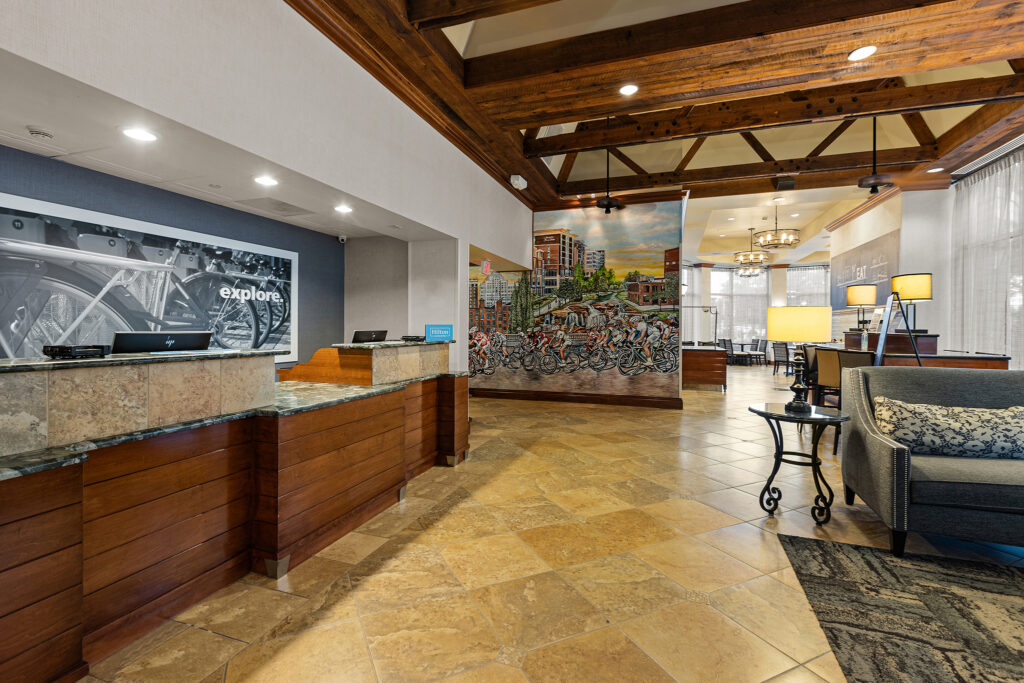 A hotel lobby with a reception desk on the left and a mural depicting a city scene with bicycles on the wall exudes the charm of Greenville. Cozy seating with lamps is on the right, while wooden beams line the ceiling above tiled floors.