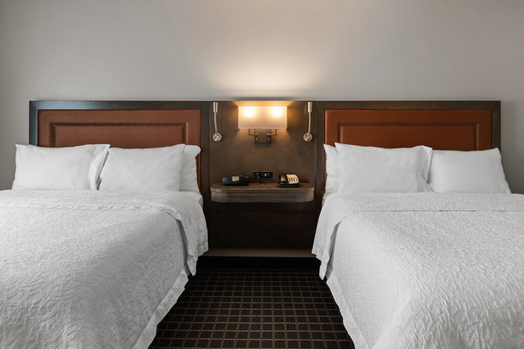 Nestled in Greenville, South Carolina, this Hilton hotel room boasts two neatly made double beds with white linens. A wooden headboard features built-in lights and a handy central shelf with a telephone and clock. The space is adorned with neutral-colored walls and a dark patterned carpet.