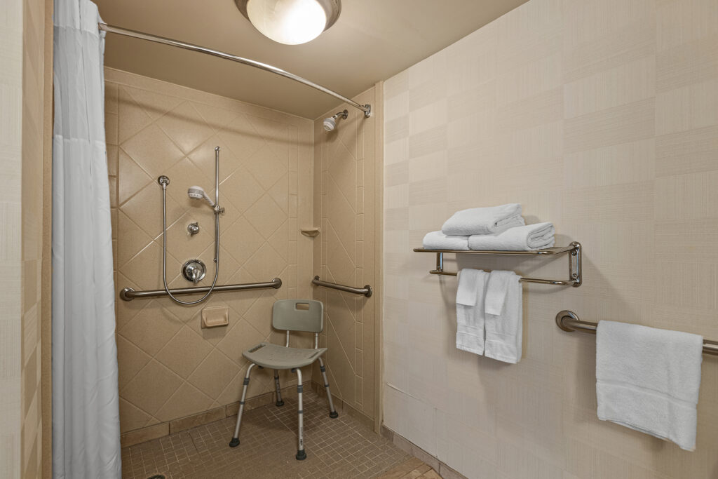 A wheelchair-accessible shower at the Hilton in Greenville, South Carolina, with grab bars, a handheld showerhead, and a shower chair. Towels are neatly stacked on a nearby metal shelf, with one towel hanging. The walls are tiled in a light color.