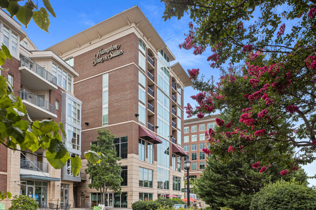 The Hampton Inn & Suites in Greenville, South Carolina, is surrounded by lush greenery and pink flowering trees. The clear blue sky perfectly complements the hotel's red brick exterior.