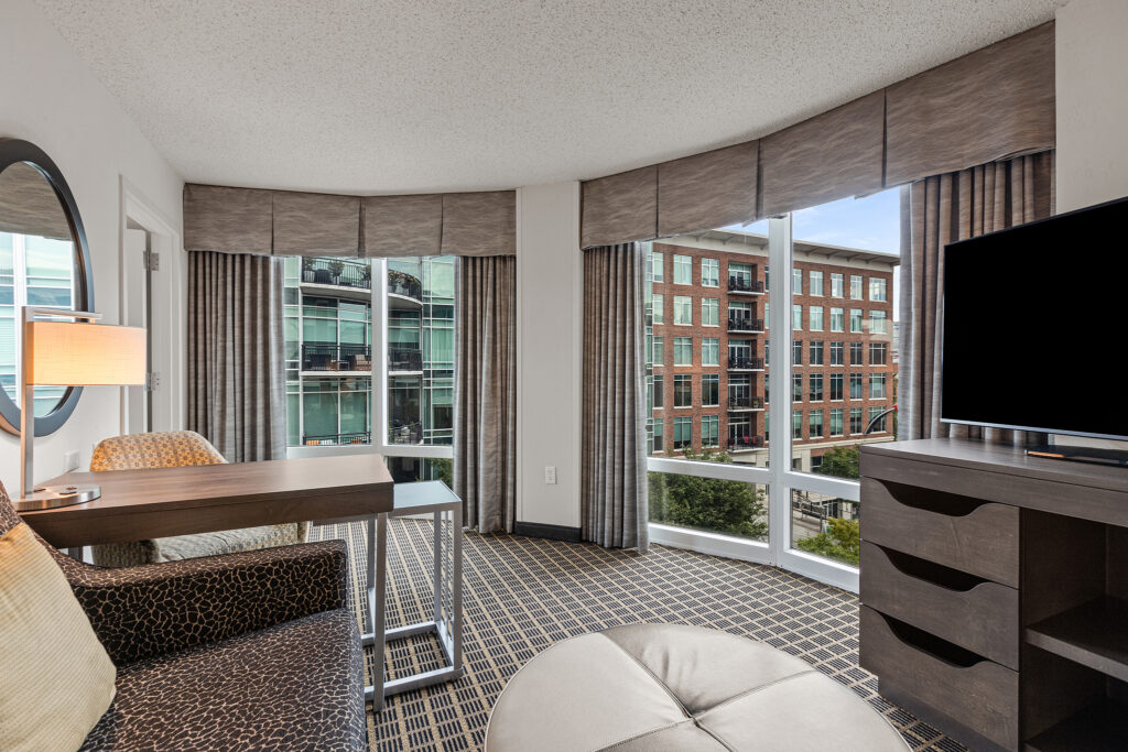 A Hilton hotel room with modern decor, featuring a desk, chair, and a large round ottoman. A flat-screen TV is on a dresser. Floor-to-ceiling windows offer views of Greenville’s urban buildings and a courtyard.