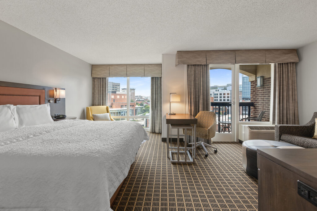 Experience a modern stay at Homewood Suites in Milwaukee. This hotel room features a large bed, armchair, desk, and chair. Bask in city views through floor-to-ceiling windows or from the balcony. Decorated in neutral tones with a woven carpet, it blends comfort and style seamlessly.