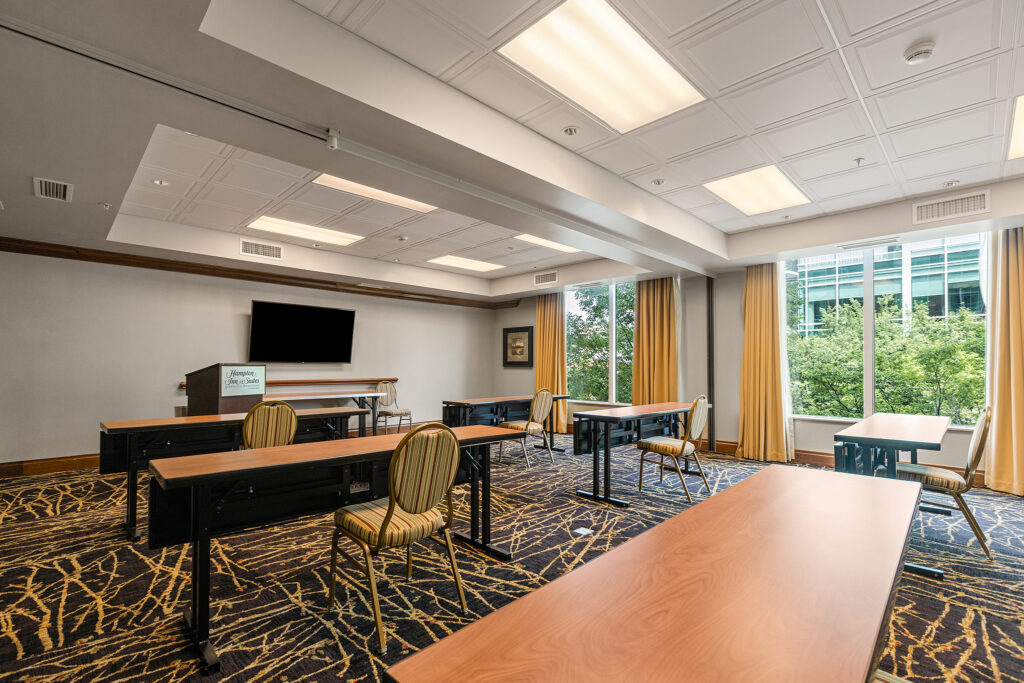 A conference room at the Greenville Hampton Inn features long rectangular tables and chairs arranged in rows. A TV is mounted on the front wall, while large windows along the side let in natural light through partially drawn beige curtains. The floor boasts a patterned carpet, enhancing its charm.