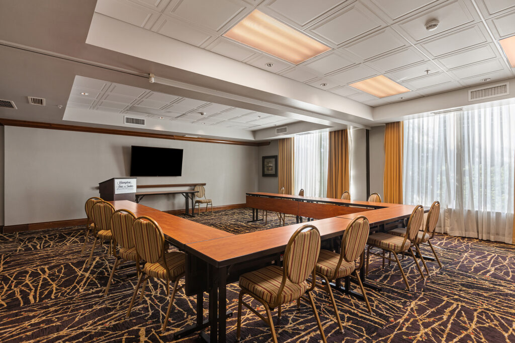 A well-lit conference room at the Hilton in Greenville, South Carolina features a U-shaped table arrangement with several chairs. A podium and large wall-mounted TV screen are at the front, complemented by patterned carpet and large windows adorned with yellow curtains.