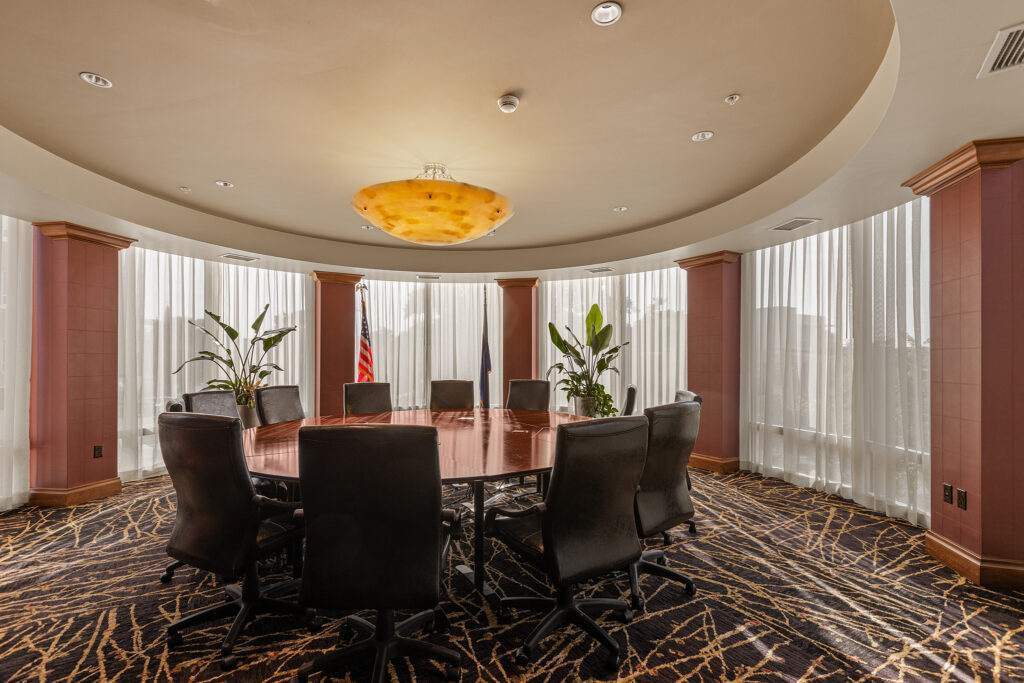 A round conference table with leather chairs sits in a well-lit room, featuring large windows with sheer curtains. An American flag is displayed proudly in the background, complemented by potted plants. This space captures the elegance of a Greenville meeting room at Hampton Inn, South Carolina.