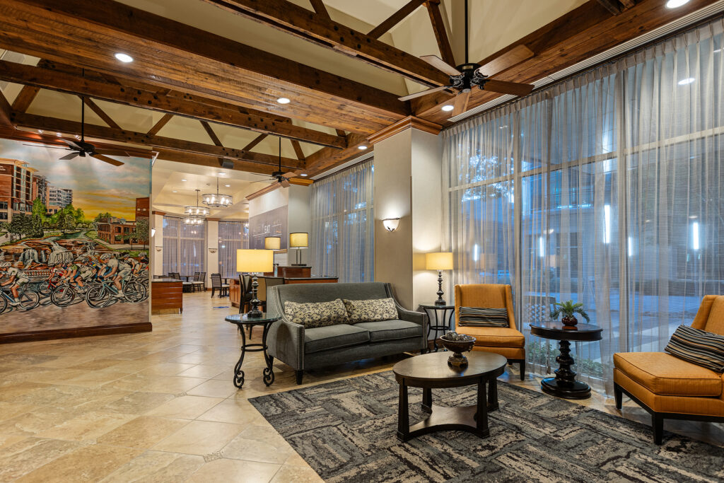 A cozy hotel lobby in South Carolina with wooden beams, soft lighting, and modern furniture. Two orange chairs and a gray sofa surround a coffee table on a patterned rug. A colorful mural and large windows with sheer curtains add to the inviting Hilton atmosphere.