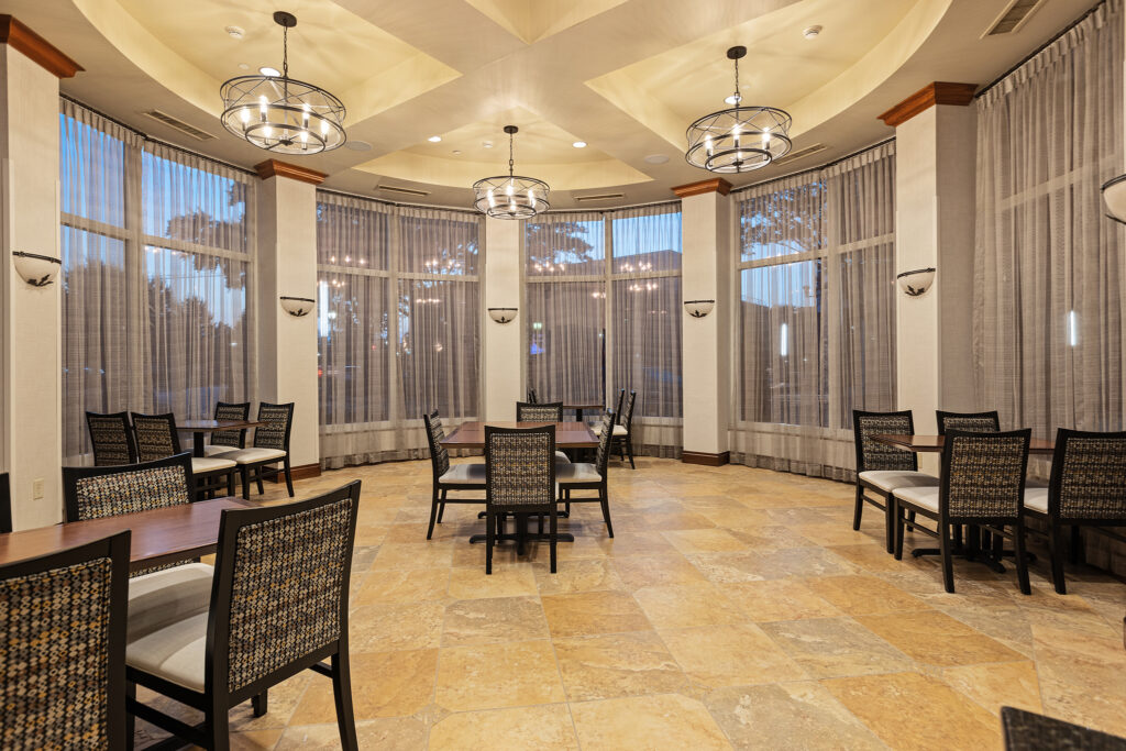 A spacious, elegant dining room in Greenville, South Carolina, features round tables and wicker chairs surrounded by tall windows with sheer curtains. Illuminated by modern chandeliers, the room's ambiance is enhanced by light stone tiled floors. Perfect for events or a cozy meal at Hampton Inn.