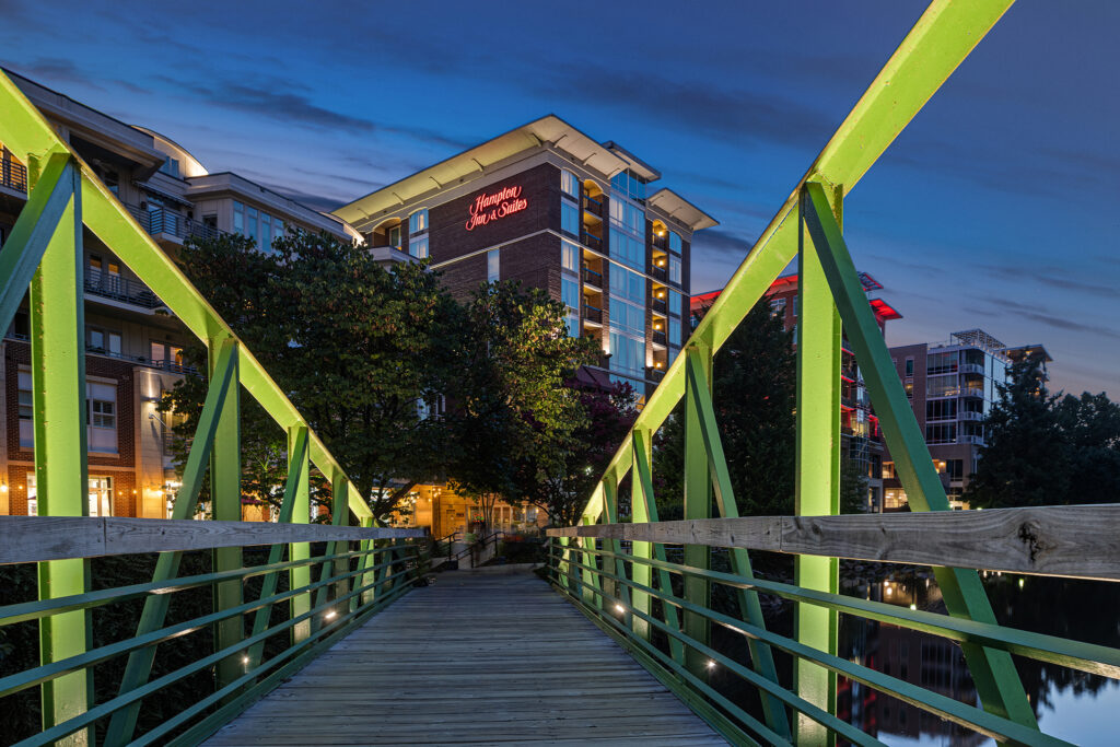 A night scene in South Carolina showcases a green pedestrian bridge leading to illuminated buildings. Trees flank the bridge, and a sign on one building reads Homewood Suites by Hilton against a deep blue sky transitioning from dusk to night.