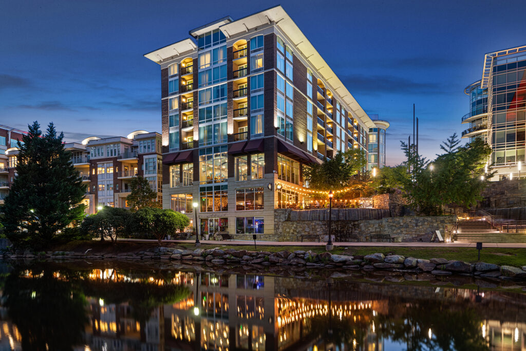 A modern, well-lit Hampton Inn by Hilton with large windows reflects in a calm body of water at dusk. String lights illuminate a terrace, and nearby trees add greenery to the urban scene under a blue evening sky, reminiscent of the charming ambiance found in Greenville, South Carolina.