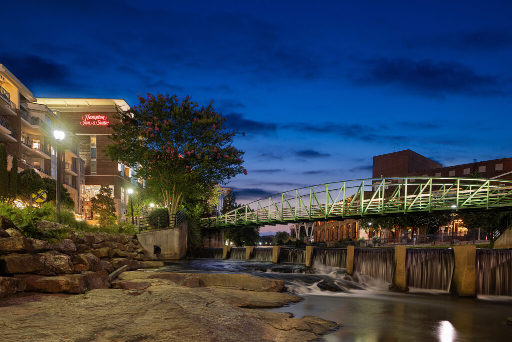 A vibrant South Carolina evening scene showcases a bridge over a gently flowing river with small waterfalls. Illuminated buildings, including a Hampton Inn & Suites, nestle amidst lush greenery, all set against the backdrop of a deep blue sky.