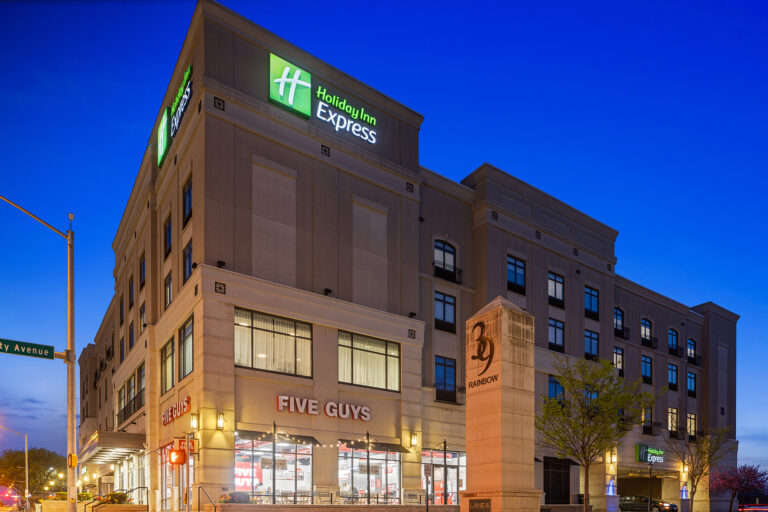 Capture the vibrant nighttime cityscape showcasing the IHG's Holiday Inn Express, a sleek multi-story hotel with Five Guys on the ground floor. The deep blue sky sets an enchanting backdrop, while a street sign anchors this perfect moment in hotel photography.