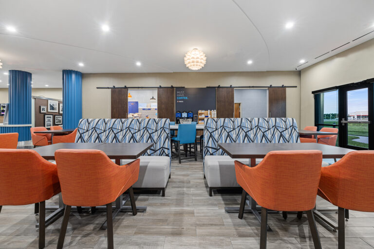 A modern hotel dining area with orange chairs around wooden tables. Behind, cushioned bench seating is paired with geometric-patterned backs. The space, perfect for hotel photography, features a coffee station and bright overhead lights, with large windows letting natural light flow in.
