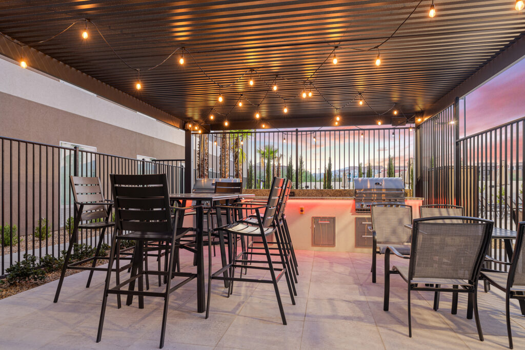 A covered outdoor patio in Las Vegas, adorned with string lights under a corrugated metal roof, features high black tables and chairs, a modern grill, and a countertop. The twilight sky and potted plants set a picturesque backdrop at this stylish Home2 Suites setting.